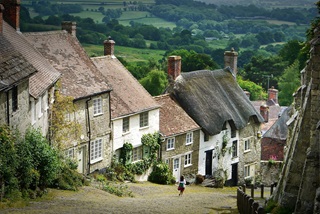 A countryside lane with houses on the left handside, looking down a hill