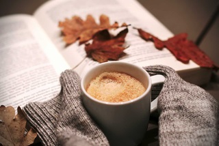 Person holding a hot drink reading a book with autumn leaves scattered on the book