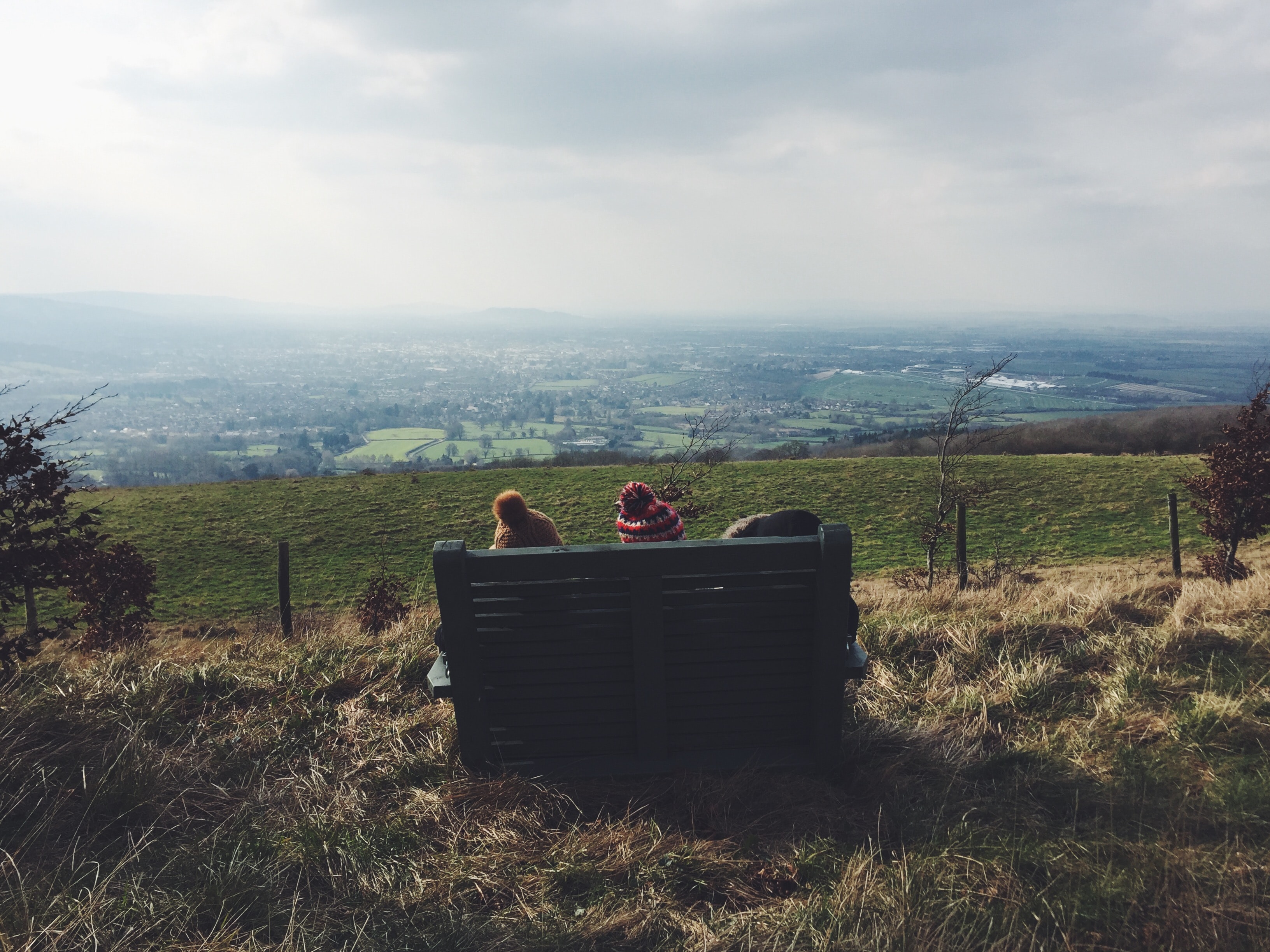 Three people wearing wolly hats sat on a bench facing a beautiful countryside view