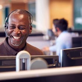 Man wearing a headset, smiling, at a computer in a call centre / telesales department