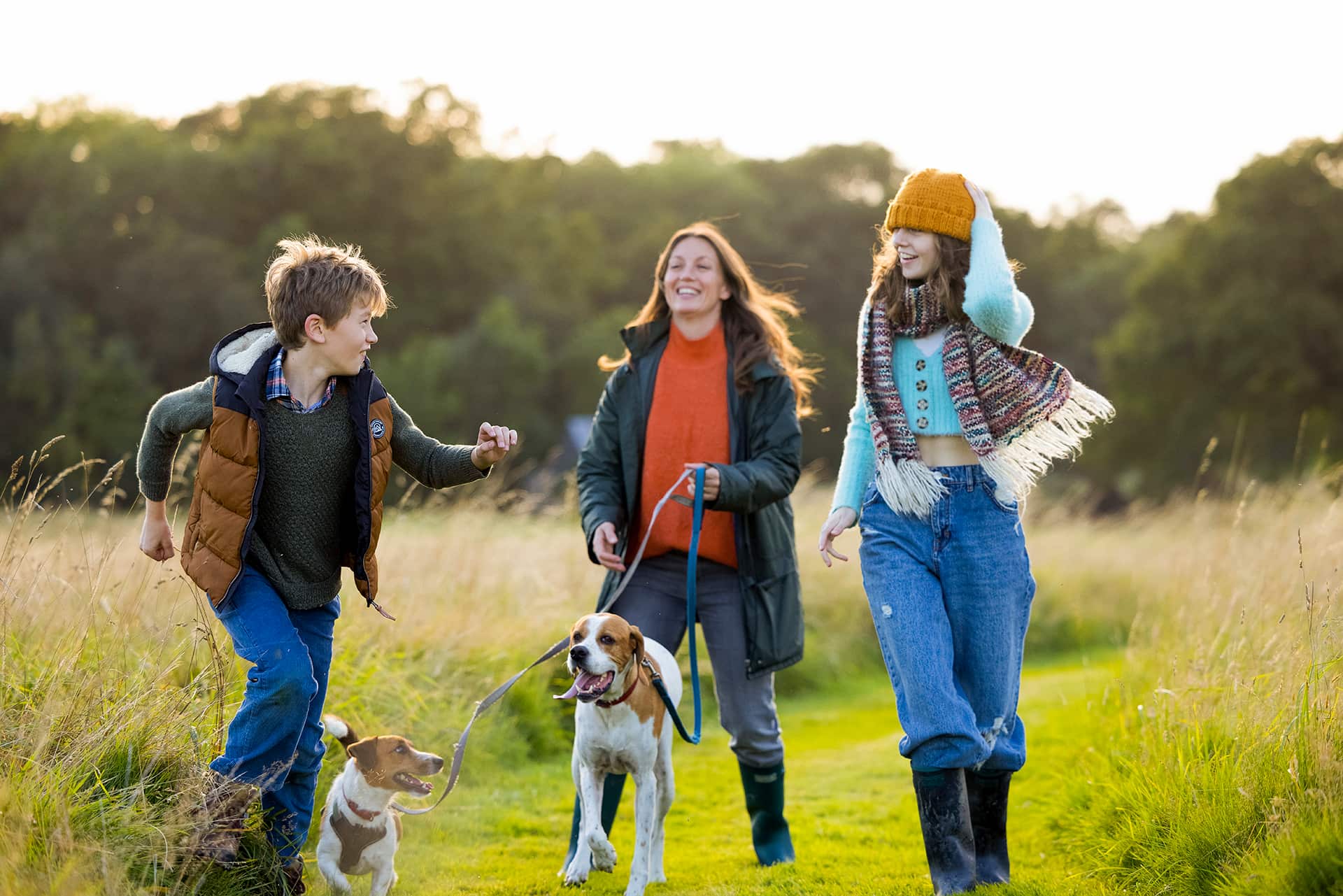 A woman, her two children, a girl and a boy and their dogs walking through a field, laughing and smiling, wearing coats and hats